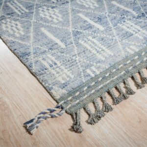 A light blue textured rug featuring geometric patterns and braided fringe rests on pale wood flooring.
