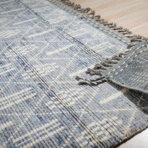A nubby blue rug with abstract white lines and fringe lays partly folded on light wood flooring.