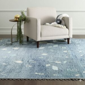 Blue textured rug with abstract white shapes and loop fringe sits under a light beige accent chair.