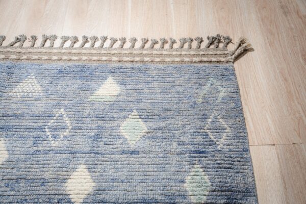 Textured blue rug with a white geometric diamond pattern, braided border, and tassels on light wood floor.