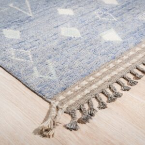 Light blue rug with white geometric designs, featuring a tasseled gray border on blonde wood flooring.