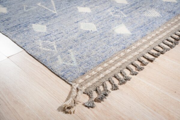 Light blue rug with white geometric designs, featuring a tasseled gray border on blonde wood flooring.
