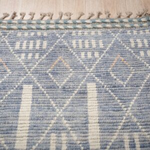 Nubby blue and cream rug with a geometric diamond pattern, braided edge, and fringe on wood flooring.