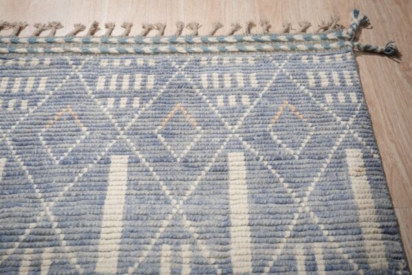 Nubby blue and cream rug with a geometric diamond pattern, braided edge, and fringe on wood flooring.