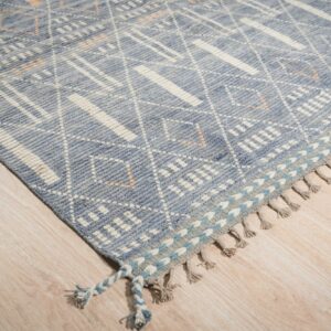 Low pile blue rug with white geometric designs and tasseled edges rests on light wood plank flooring.