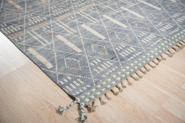 Low pile blue rug with white geometric designs and tasseled edges rests on light wood plank flooring.