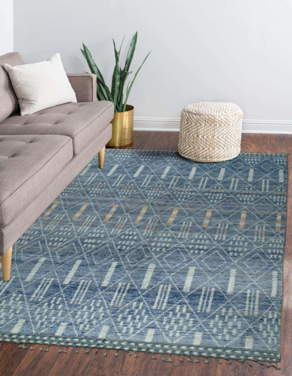 Blue geometric pattern rug with white and orange accents sits on a wood floor next to a gray sofa and braided pouf.