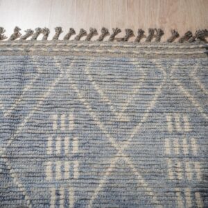 Textured blue and cream rug featuring a geometric pattern, thick fringe, and light wood flooring backdrop.