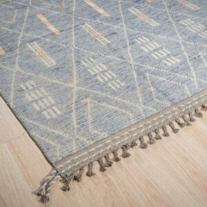 Blue rug with white geometric designs and orange accents has a braided grey tasseled border.