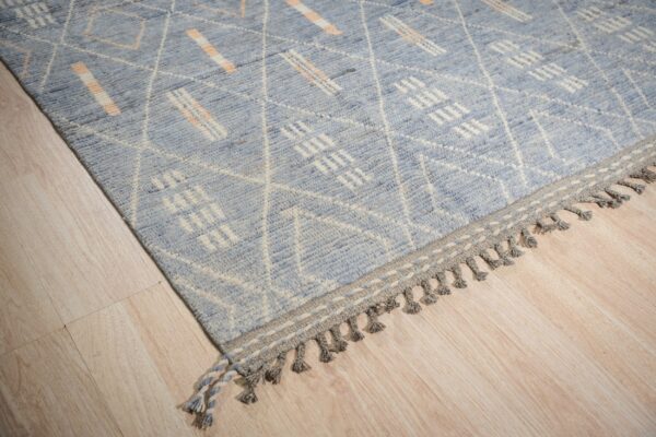 Blue rug with white geometric designs and orange accents has a braided grey tasseled border.