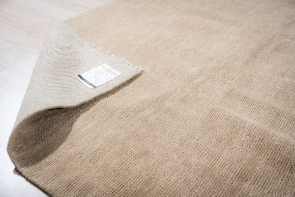 Light tan, high-pile rug with ribbed texture, partially rolled up on pale wood planks.