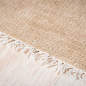 A light beige high-pile textured rug edge with white fringe lies on pale wood planks.