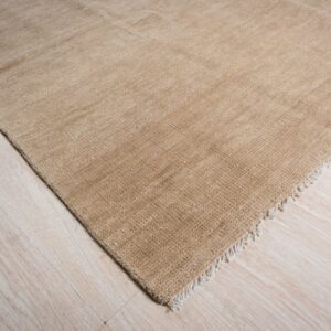 Textured, solid tan rug with a short fringe lying on light blonde wood flooring.