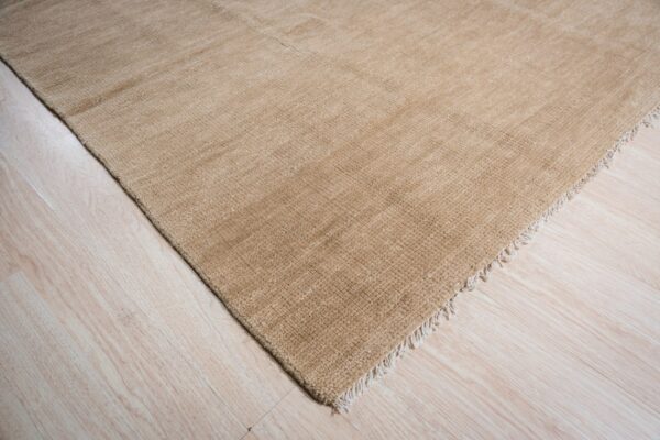 Textured, solid tan rug with a short fringe lying on light blonde wood flooring.