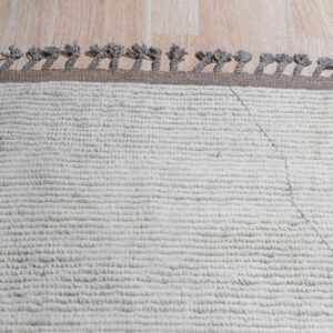 Close-up of a high-pile white rug featuring a brown border and gray twisted tassels on wood flooring.