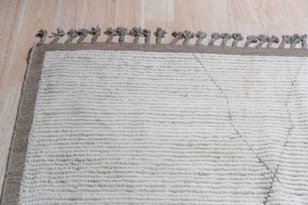 Close-up of a high-pile white rug featuring a brown border and gray twisted tassels on wood flooring.