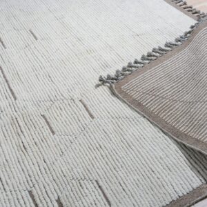 Off-white textured rug with geometric brown lines lays on wood floor, showing a striped, tasseled backing.