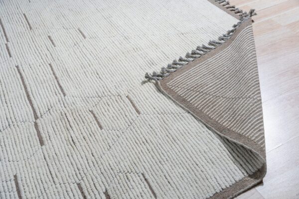 Off-white textured rug with geometric brown lines lays on wood floor, showing a striped, tasseled backing.
