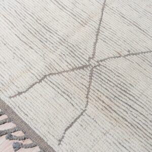 High-pile cream rug with grey geometric lines, parallel ribbing, and thick grey tassels resting on light wood.