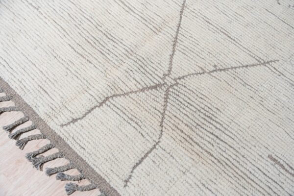 High-pile cream rug with grey geometric lines, parallel ribbing, and thick grey tassels resting on light wood.