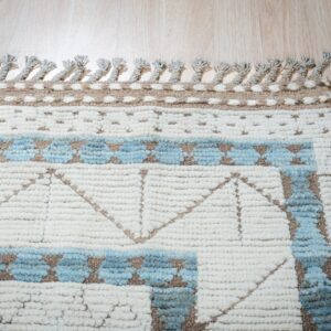 Textured cream rug with light blue and brown geometric patterns and knotted tassels lying on wood flooring.