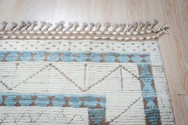 Textured cream rug with light blue and brown geometric patterns and knotted tassels lying on wood flooring.
