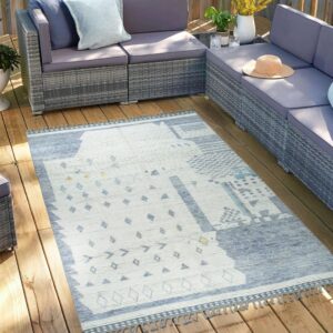 Geometric blue and white rug with tassels on a sunlit wooden deck, next to grey patio furniture.