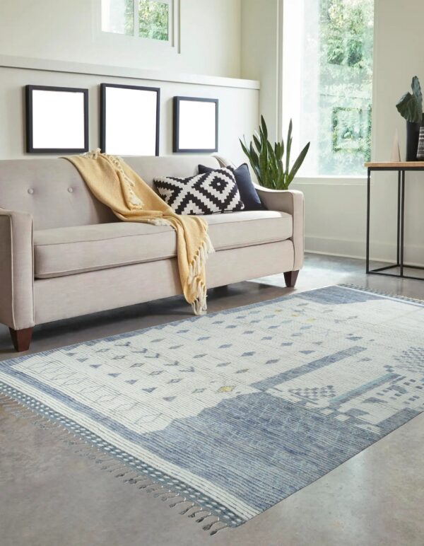 Abstract blue and white low-pile rug with fringe accents beneath a beige sofa on gray flooring.