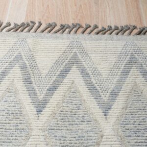 A high-texture cream and grey rug with a geometric pattern and twisted fringe rests on wood plank flooring.