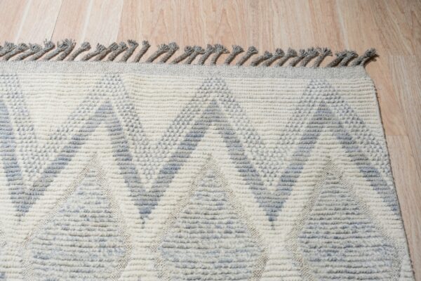 A high-texture cream and grey rug with a geometric pattern and twisted fringe rests on wood plank flooring.