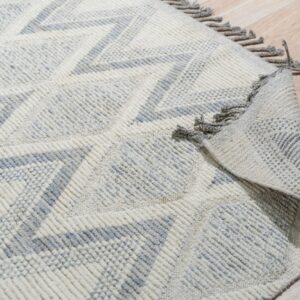 Cream and gray textured geometric rug with fringe, partially rolled on light wood flooring.