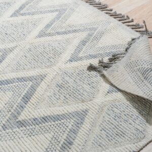 Textured off-white and light gray rug with geometric zigzags and tassels lying on wood plank flooring.