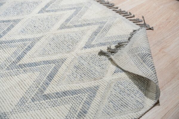 Textured off-white and light gray rug with geometric zigzags and tassels lying on wood plank flooring.