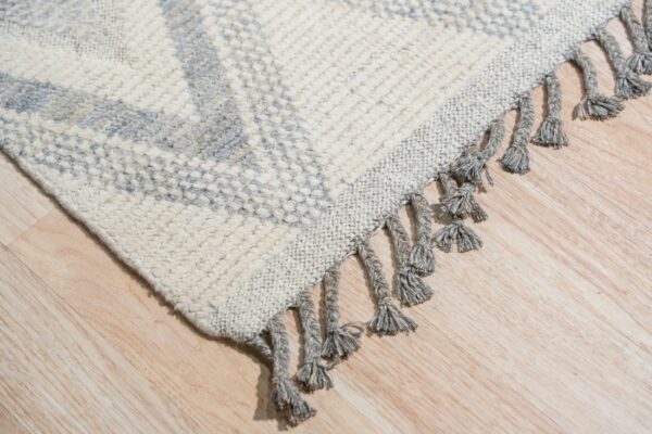 Textured cream rug corner featuring a light blue geometric pattern and thick gray tassels on wood flooring.