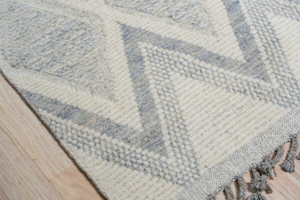 Geometric cream and pale blue textured rug detail showing knotted fringes on light hardwood.