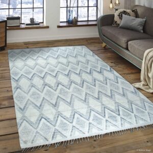 White and blue zigzag patterned rug on wood floors, next to a gray sofa near industrial windows.