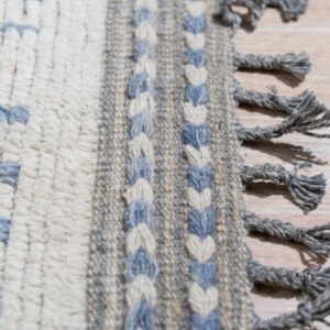 Close up of a cream rug with blue geometric shapes, braided borders, thick gray fringe, lying on light wood.
