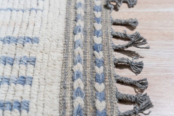 Close up of a cream rug with blue geometric shapes, braided borders, thick gray fringe, lying on light wood.