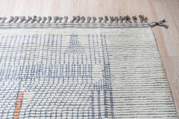 Textured cream rug with blue grid pattern, a visible orange stripe, and gray knotted fringe on wood floor.