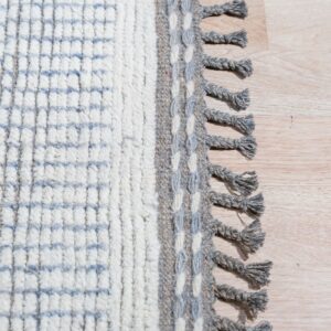 High-pile white patterned rug with a gray woven border and braided tassels on light wood flooring.