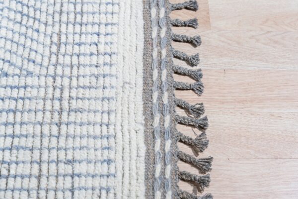 High-pile white patterned rug with a gray woven border and braided tassels on light wood flooring.