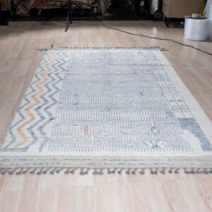 White rug with blue and orange geometric patterns and gray tassels lying on light wood plank flooring.
