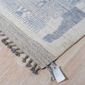 Textured ivory and light blue rug with gray braided edges and tassels rests on light wood floor.