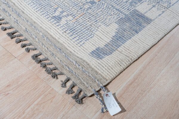 Textured ivory and light blue rug with gray braided edges and tassels rests on light wood floor.