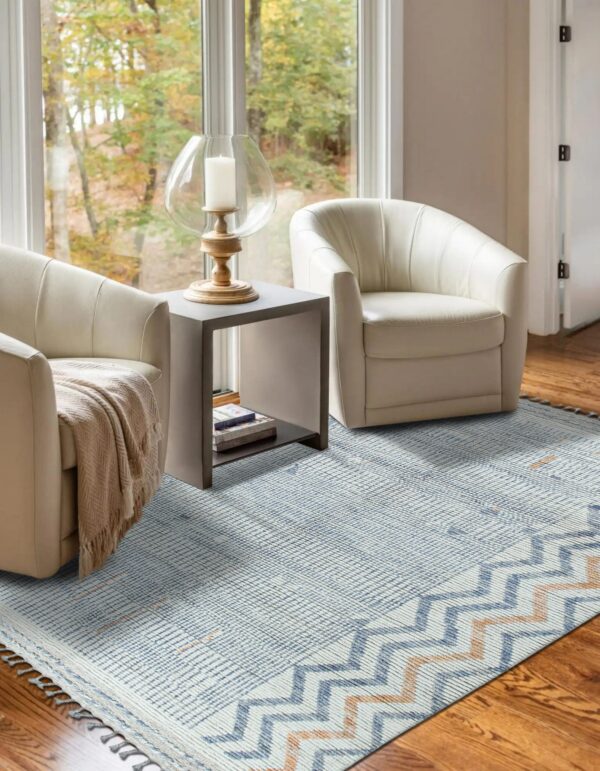Blue and cream flatweave rug with chevron borders between two white chairs on a hardwood floor by a window.