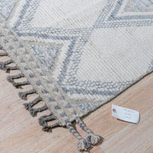 Chunky knit rug featuring cream and light gray geometric diamonds and thick gray tassels rests on wood planks.