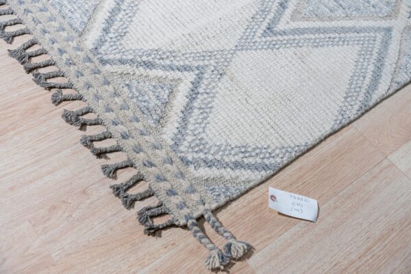 Chunky knit rug featuring cream and light gray geometric diamonds and thick gray tassels rests on wood planks.
