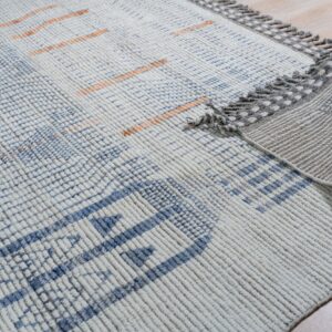 Chunky textured white rug with blue and subtle orange geometric patterns and decorative gray tassels on wood floor.