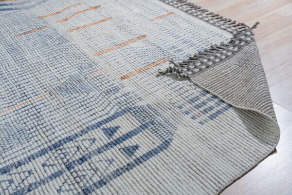 Chunky textured white rug with blue and subtle orange geometric patterns and decorative gray tassels on wood floor.