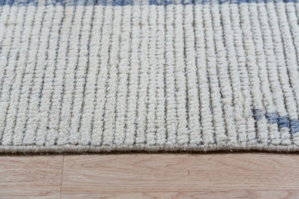 Close view of a cream ribbed rug featuring thin vertical dark threads and blue pattern accents on a wood floor.